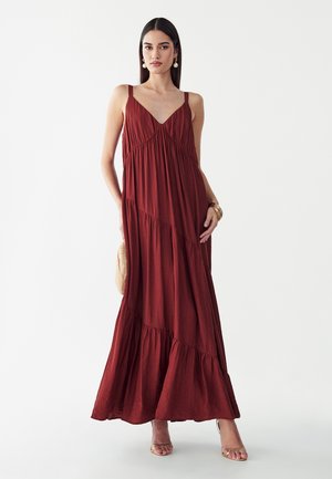 Mujer con un vestido largo sin mangas de color rojo intenso con escote en V, sosteniendo un clutch tejido, con accesorios de pendientes y pulseras doradas.