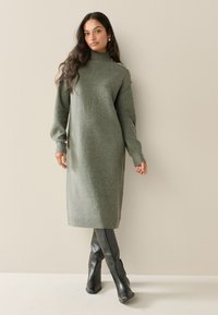Vestito lungo verde oliva lavorato a maglia con collo alto, maniche a coste e lievi decorazioni sulle spalle, abbinato a stivali neri al ginocchio.