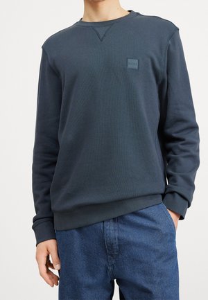 Homme portant un sweat-shirt à col rond bleu marine avec un petit patch logo "BOSS" et un pantalon bleu texturé, mains relaxées le long du corps.