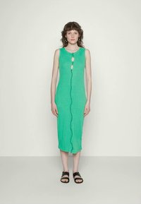 Mulher com cabelo castanho encaracolado, vestindo um vestido midi verde sem mangas com detalhes recortados e sandálias pretas, de pé contra um fundo simples.