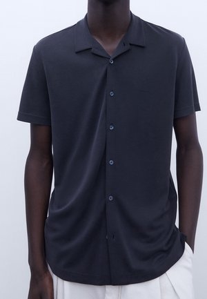 Homme portant une chemise noire à manches courtes avec un col camp et un pantalon blanc, debout devant un fond uni.