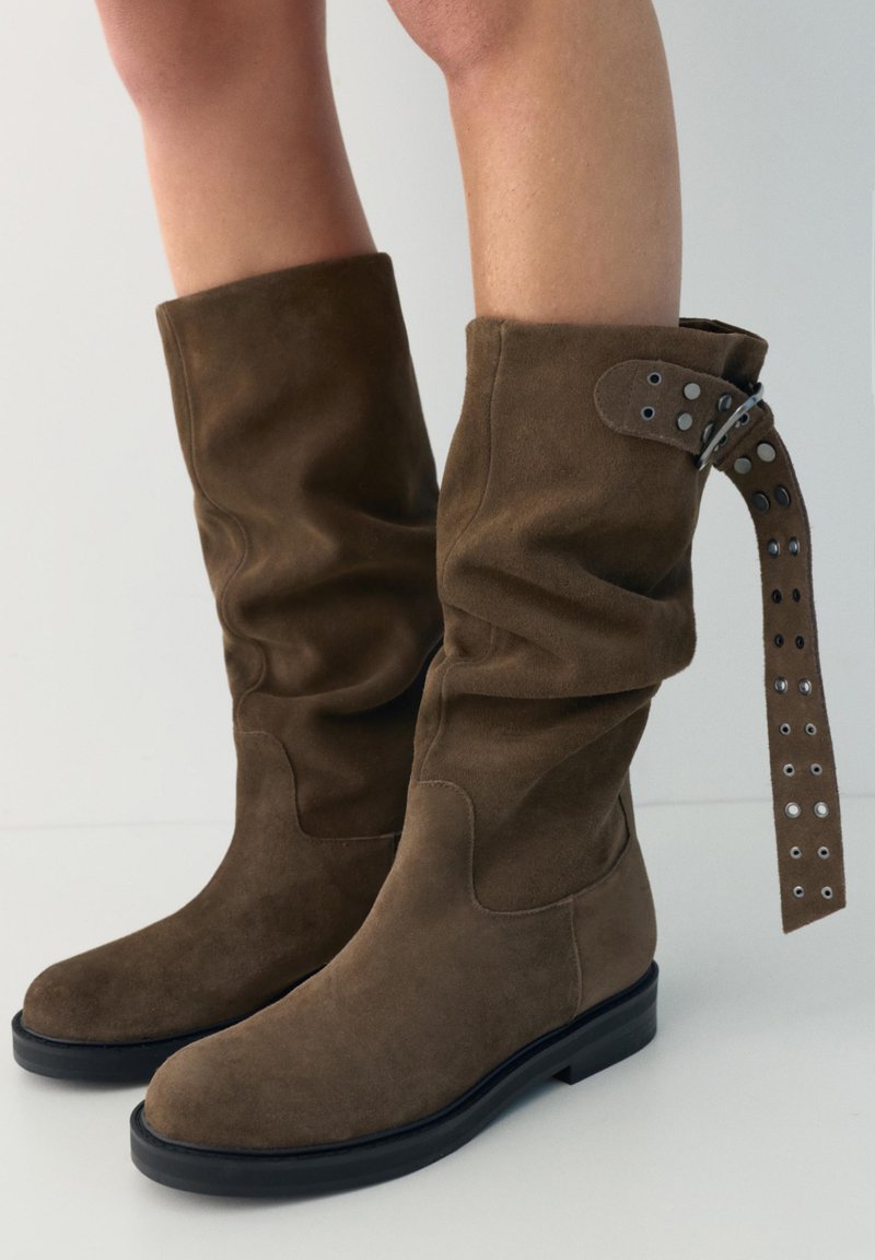 Bottes en daim marron mi-mollet avec semelles noires, coupe ample et une sangle allongée avec œillets métalliques enroulée autour de la tige de la botte droite.