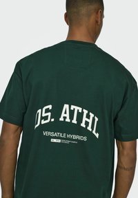 Tamsiai žalia medvilnės marškinėliai su balta spauda, kurioje užrašyta „OS. ATHL“ ir „VERSATILE HYBRIDS“ ant nugaros. Trumpomis rankovėmis, apvaliu kaklu.