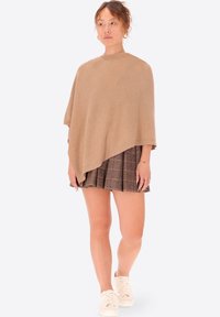 Poncho beige realizzato in tessuto morbido, caratterizzato da un design semplice e asimmetrico. Abbinato a una mini gonna a quadri e scarpe da ginnastica bianche.
