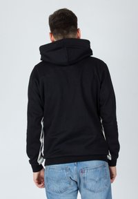 Hombre con sudadera negra y jeans azules de pie de espaldas contra un fondo gris claro liso.