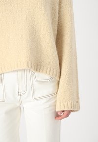 Beige gebreide trui met een gestructureerde afwerking, relaxed fit en geribbelde manchetten, gecombineerd met witte broek met contrasterende stikseldetails.