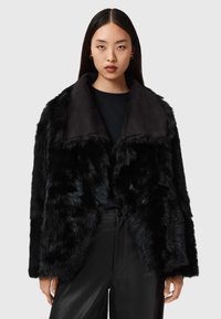 Giacca in pelliccia sintetica nera con ampio colletto e tessuto liscio. Presenta un esterno testurizzato e soffice e un design elegante, abbinata a pantaloni neri.