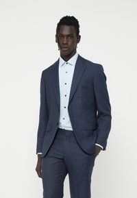 Sacou elegant bleumarin, cu revere tăiate, asortat cu o cămașă de culoare bleu deschis, având accente de nasturi închise la culoare. Textura fină a materialului.