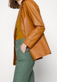 Tan leren jack met een ontspannen pasvorm, gedragen over een mosterdkleurig topje en groen geruite broeken. Opvallende knoopdetails op het jack.