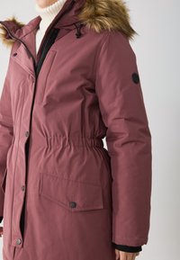 Bordový parka s kapucňou podšitou kožušinou, elastickým pásom, zipsom a patentkami, a prednými vreckami. Obsahuje hladký, lesklý materiál.