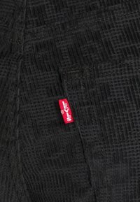 Černá texturovaná tkanina s mřížkovým vzorem. Na levé straně se nachází červená nášivka Levi's, která ukazuje detaily šití a prošívání.