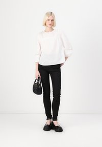 Bílá, texturovaná blůza s krajkovými detaily, černé skinny džíny a černá kabelka s tkaným vzorem. Modelka stojí na jednobarevném pozadí.