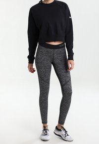 Felpa nera a maniche lunghe con lettering testurizzato e leggings grigi a fantasia. Il modello indossa sneakers nere, mostrando un look sportivo.