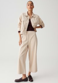Vrouw in een beige geribde jas en wijde broek over een donkerbruin top, gecombineerd met donkerbruine leren loafers, staande tegen een witte achtergrond.