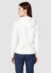 Sudadera blanca de algodón con capucha ajustable, puños y dobladillo acanalados, mostrada desde la parte posterior, combinada con jeans de mezclilla oscura.