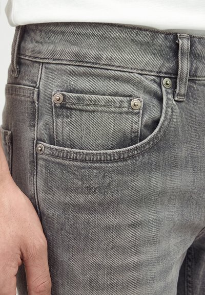 Gros plan d'une poche de jean en denim gris avec rivets et une main reposant à côté, montrant la texture et les détails des coutures.