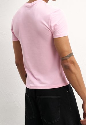 Homme portant une chemise rose clair moulante à manches courtes et un pantalon noir, vu de dos avec un tatouage à motifs sur son bras droit.