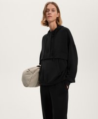 Felpa nera oversized con tasca a marsupio, cappuccio con cordino e texture liscia. Una borsa beige imbottita è tenuta di lato.