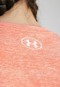 Primer plano del cuello y la parte superior de la espalda de una persona que lleva una camiseta coral-pink de Under Armour con el logotipo centrado debajo del escote.