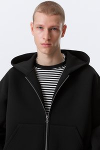 Jeune homme aux cheveux courts portant un sweat à capuche noir zippé sur un t-shirt rayé noir et blanc et un collier chaîne argenté.