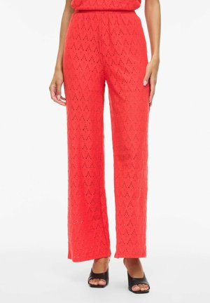 Czerwone szerokie spodnie z wzorem z koronki w kształcie diamentu, noszone z czarnymi sandałami na obcasie z odkrytymi palcami, ręce opierające się przy bokach.