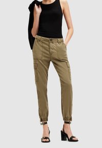 Femme portant un pantalon cargo vert olive avec des poignets élastiques, un débardeur noir, une veste noire sur l'épaule et des sandales à talons noires.