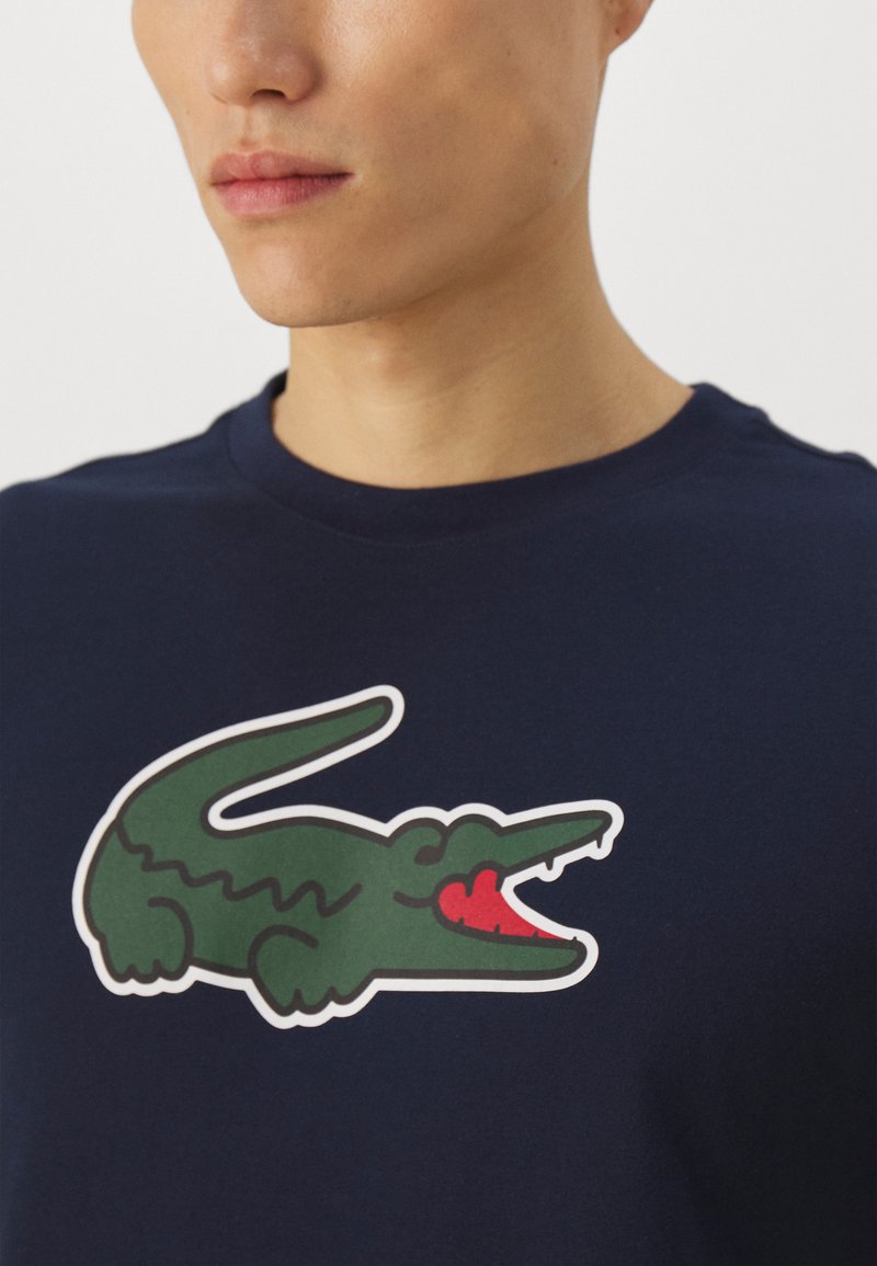 T-shirt blu navy con logo di coccodrillo verde e dettagli rossi. Il design ha una texture liscia e un classico scollo rotondo.