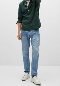 Man wearing light blue jeans, white sneakers, a dark green button-up shirt over a white t-shirt, standing with one arm raised.