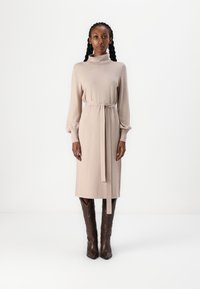 Abito beige lavorato a maglia con colletto alto, maniche lunghe e una cintura da legare in vita. L'abito presenta polsini a sbuffo e scende sotto il ginocchio.
