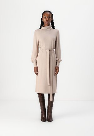 Beige neulemekko, jossa on korkeakauluksinen malli, pitkät hihat ja vyötäröllä solmittava vyö. Mekon hihansuissa on puffiyksityiskohtia, ja se ulottuu polvien alapuolelle.