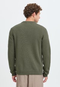Mann mit lockigem Haar trägt einen olivgrünen Pullover und beige Hosen, steht mit dem Rücken zur Kamera vor einem schlichten hellen Hintergrund.