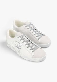 Zapatillas de cuero blancas con detalles en ante gris, cordones plateados con un patrón y un logo de calavera y tibias cruzadas en el lateral. Textura suave.