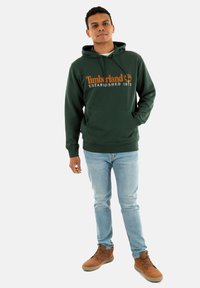 Sudadera verde oscura con el logo y el texto naranja de Timberland; bolsillo canguro; combinada con jeans azul claro y botas marrones con cordones.