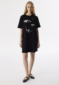 Zwarte t-shirt jurk met korte mouwen, met abstracte witte illustraties en tekst op de voorkant. Zachte katoenen stof, ontspannen pasvorm.