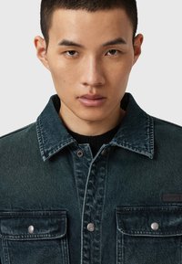 Tummansininen farkkutakki, jossa on kaksi etutaskua, hopeiset napsunapit ja terävä kaulus, jossa on näkyvät ompelu yksityiskohdat.