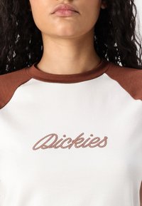 Hvit T-skjorte med brune raglan-ermer. Har "Dickies" brodert i tau-stil. Slank tekstur med rund hals.
