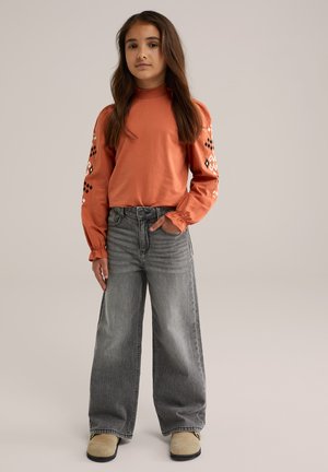 Oranges Langarmhemd mit Rüschensaum und gemusterten Ärmeln, kombiniert mit grauen High-Waist-Jeans mit weitem Bein und beigen Slippern.
