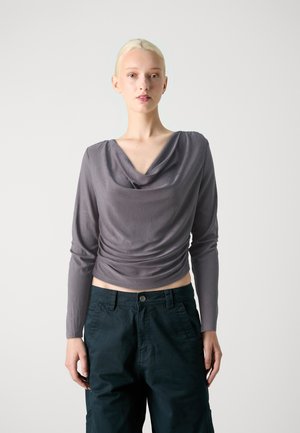 Haut long gris à manches avec un col drapé, présentant un corsage ajusté et un détail froncé sur le côté, associé à un pantalon vert foncé.