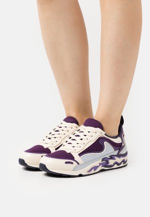Jambes portant des baskets à lacets violettes et crème avec des panneaux en mesh et des semelles à motif ondulé sur un fond blanc uni.