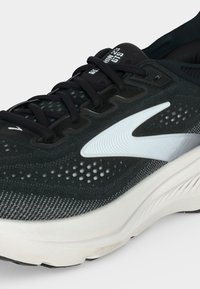 Chaussure de course noire et blanche avec lacets noirs, tige en tissu perforé et semelle blanche épaisse inclinée pour montrer le logo latéral et le texte sur la languette.