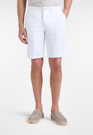 Uomo con pantaloncini bianchi fino al ginocchio, mocassini grigio chiaro e camicia chiara infilata, in piedi con una mano in tasca.