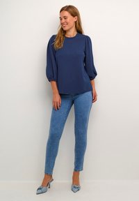 Blusa blu navy con maniche a sbuffo, abbinata a jeans skinny blu chiaro e tacchi decorati con fiocco blu. Tessuto liscio con una sottile texture.