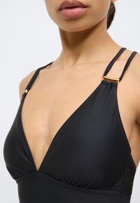 Juodas trikampio formos bikini viršus su plonomis petnešėlėmis ir auksiniais akcentais; pasižymi lygiu audiniu ir raukiniais krūtinės srityje.