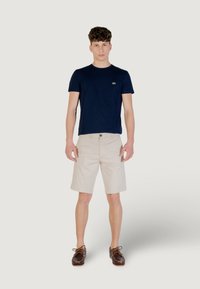 T-shirt en coton bleu marine avec col rond, shorts beiges et chaussures en cuir marron avec lacets. Modèle se tenant contre un fond neutre.