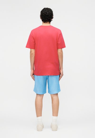 Άνδρας με σγουρά σκουρόχρωμα μαλλιά, φοράει απλό κόκκινο μπλουζάκι, ανοιχτό μπλε σορτς, λευκές κάλτσες και εκρού αθλητικά παπούτσια, στέκεται γυρισμένος με την πλάτη.