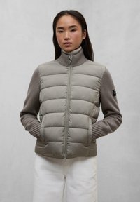 Gilet imbottito in grigio chiaro con colletto alto e maniche a maglia. Presenta una zip frontale, tasche laterali e polsini a coste.