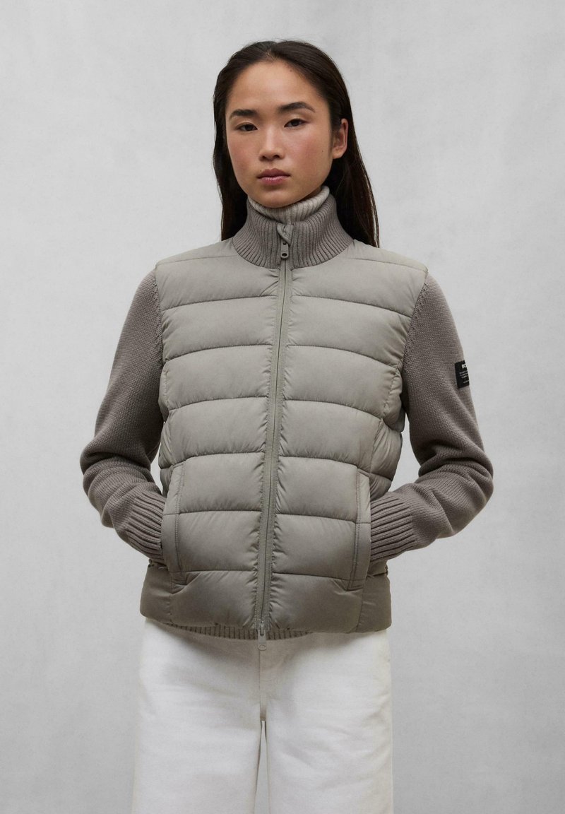 Gilet imbottito in grigio chiaro con colletto alto e maniche a maglia. Presenta una zip frontale, tasche laterali e polsini a coste.