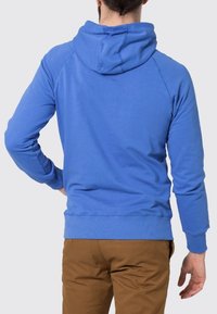 Sudadera de algodón azul con un bolsillo canguro, capucha ajustable con cordón y puños y dobladillo acanalados. Usada con pantalones marrones.