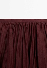 Tissu bordeaux profond rassemblé en plis serrés sous une large ceinture, montrant la texture et les plis d'un matériau doux et léger.