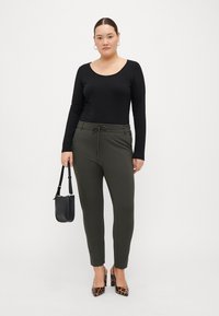 Top negro de manga larga, pantalones de tiro ajustado verde oliva con cintura con cordón, y tacones de leopardo; bolso negro sostenido con una mano.
